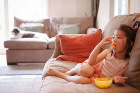 A pretty little girl is watching TV while eating cheese puffs from a living room in her home. She's alone with her pet dog, who is lying on the couch in the background. She's 6 years old and is of Eurasian decent.